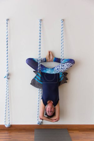Foto de Yoga teacher hanging upside down on ropes from white wall in studio. Yogi as bat like posture stretching whole body. Flexibility practice, healthy lifestyle concept - Imagen libre de derechos