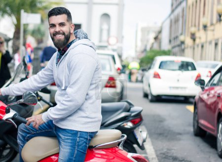 Foto de Feliz joven estacionando ciclomotor rojo en el casco antiguo de Las Palmas, España. hipster barbudo completo sentado en la clásica motocicleta italiana de estilo retro en la calle - Imagen libre de derechos