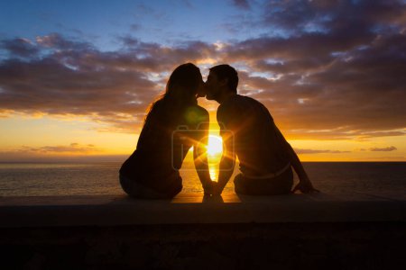 Téléchargez les photos : Man and woman sitting by the sea kiss at sunset at Meloneras beach walk, Gran Canaria. Couple silhouette enjoying colorful twilight. Valentines Day, honeymoon romantic date concepts - en image libre de droit