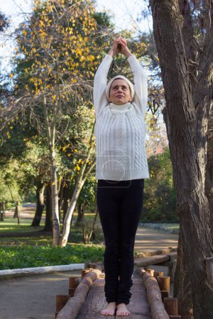 Photo for Mature woman on wooden path wearing warm clothes practices yoga early morning in the park. Middle aged lady on mountain pose with extended arms overhead as part of sun salutation - Royalty Free Image