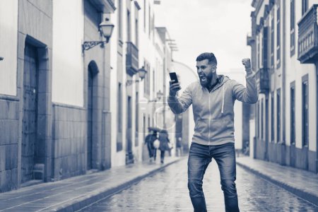 Photo for Lucky young man with fist up celebrates victory holding cellphone on rainy street in the city. Full beard hipster model on grey hoodie with euphoric expression. On line bet winner, exultant concept - Royalty Free Image