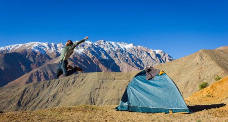 Foto de Big jump of energetic young man next to igloo camping tent with magnificent views of Andes snowed mountain range on the background under blue sky in Elqui Valley, Chile. Explore, hiking concepts - Imagen libre de derechos