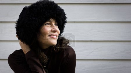 Photo for Smiling mature lady holds black fur Russian hat on head over white wood plank wall background. Middle age pretty woman happy on cold weather. Ready for winter season. Female fashion, trendy concepts - Royalty Free Image