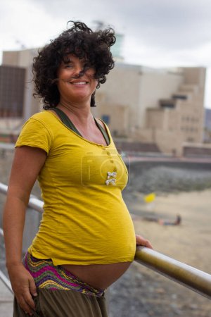 Foto de Feliz madre embarazada en el día ventoso junto al mar. Jovencita sonriente con camisa amarilla esperando un bebé en la barandilla de playa en Las Palmas, España. Salud de la mujer, bienestar - Imagen libre de derechos