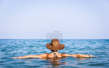 Foto de Young sexy woman facing away towards horizon with opens arms on water surface wearing wicker pamela hat and bikini on the beach in the island of Koh Phangan, Thailand. Relax, peaceful vacation concept - Imagen libre de derechos