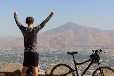 Photo for Young man with arms up next to bike with views of Santiago city on sunny day. Triumphant cyclist on top of mountain in capital of Chile - Royalty Free Image