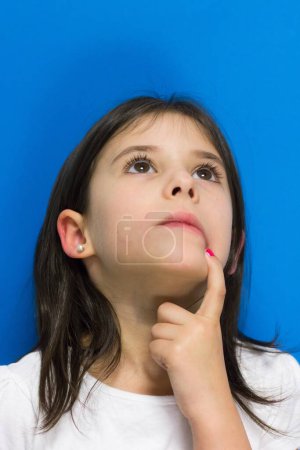 Foto de Little girl looking up with finger on mouth thinking of an idea over blue background. Imagination, vision, inspiration concept - Imagen libre de derechos