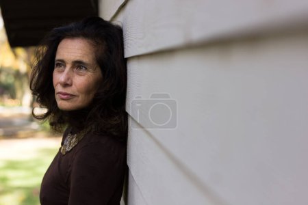 Téléchargez les photos : Serious mature woman on brown sweater and necklace leaning against white wooden plank wall. Middle aged classy lady with concerned, worried look. Retirement, menopause, problem concepts - en image libre de droit