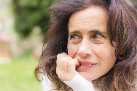 Foto de Portrait of lovely mature lady with thoughtful dreamy look while bites her nails in the park. Middle aged beautiful woman project imagination idea concept - Imagen libre de derechos