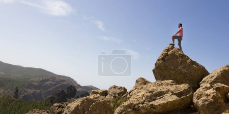 Photo for Young man standing on rock top on sunny day in Roque Nublo national park, Gran Canaria. Hiker observing natural landscape panoramic mountain view in Canary Islands, Spain. Explorer, visionary concepts - Royalty Free Image