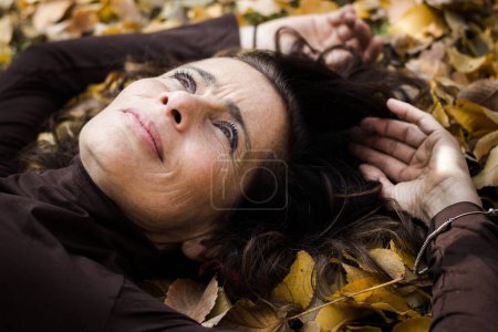 Photo for Glamorous mature woman lies on brown leaves on the ground with hands overhead and thoughtful look. Middle aged pretty lady with hopeful, dreamy vision. Female model fall season fashion sales concept - Royalty Free Image