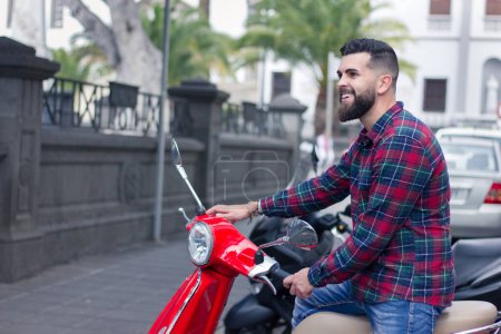 Foto de Joven hipster con sonrisa sentado en scooter rojo en la ciudad. Hermoso hombre barbudo completo estacionamiento motocicleta en la calle. Modelo masculino en vaqueros azules y camisa de franela de damas rojas - Imagen libre de derechos