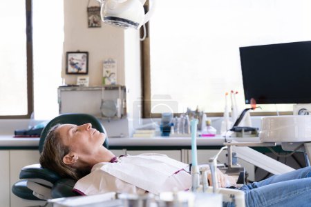 Photo for Middle aged woman on dentist stretcher with tools all over the room. Female patient with eyes closed waiting for checkup - Royalty Free Image