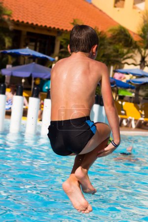 Téléchargez les photos : Young boy diving into swimming pool. Kid holding legs in the air jumping into water. Summer vacation, resort fun holidays, leisure time concepts - en image libre de droit
