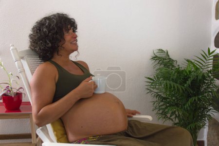Foto de Mujer embarazada sonriendo en mecedora con taza de café blanco en el vientre. Feliz madre embarazada descansando en la sala de estar en casa - Imagen libre de derechos