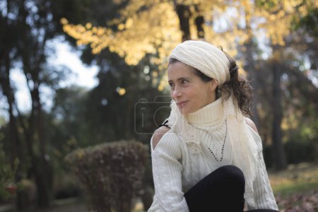 Téléchargez les photos : Hippie mature woman portrait. Lovely middle aged lady posing with white turtle neck sweater and shawl on head in the park. Autumn fashion model, fall sales, healthy lifestyle, young looking concepts - en image libre de droit