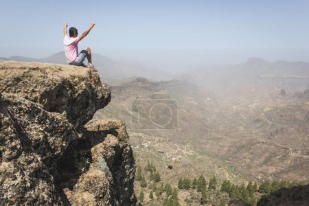 Photo for Fearless hiker sitting on top of rock edge enjoying mountain landscape in Gran Canaria. Confident man celebrating nature beauty with arms up. Courage, dare, no fear, victory, winner concepts - Royalty Free Image