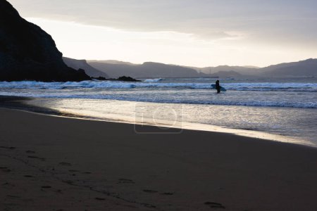 Téléchargez les photos : Silhouette de femme entrant dans l'eau avec planche de surf au coucher du soleil sur la plage vide d'Atxabiribil, Pays Basque. Passion pour le surf en hiver concept - en image libre de droit
