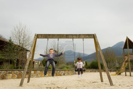 Téléchargez les photos : Deux gamins se balançant à l'aire de jeux et garçon sautant de balançoire par temps nuageux. Concept amusant pour l'enfance - en image libre de droit