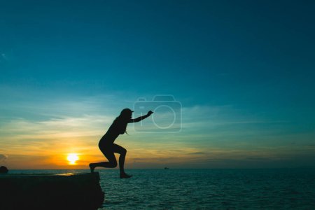 Silhouette of brave girl jumping off cliff to ocean at splendid sunset in the island of Ko Phangan, Thailand. Dare, no fear concept