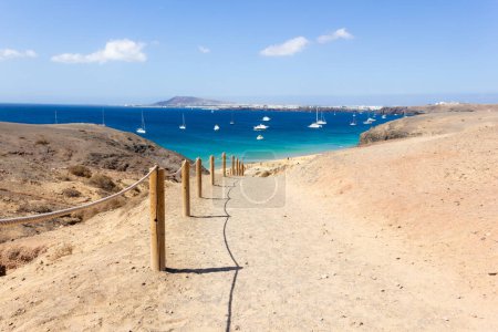 Téléchargez les photos : Chemin de terre menant à la plage de Papagayo avec voiliers sur l'eau de mer turquoise sur ensoleillé à Lanazarote. Vacances d'été, concepts de destination de voyage - en image libre de droit