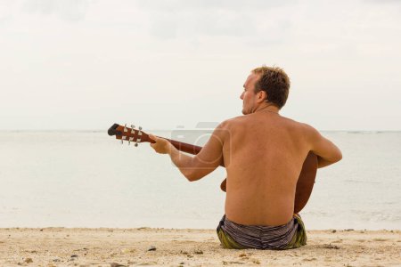Téléchargez les photos : Jeune homme blond en maillot de bain assis sur la plage jouant de la guitare tout en cherchant l'inspiration dans l'île de Koh Phangan, Thaïlande. Hippie profiter de la solitude pour composer - en image libre de droit