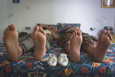 Photo for Mom and dad barefoot lying in bed with white baby shoes in the middle. Feet of happy parents expecting baby in their bedroom. Parenthood, new life concept - Royalty Free Image