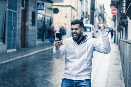 Téléchargez les photos : Lucky young man with fist up celebrates victory holding cellphone on rainy street in the city. Full beard hipster model on grey hoodie with euphoric expression. On line bet winner, exultant concept - en image libre de droit
