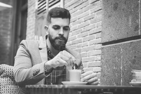 Photo for Elegant businessman having cup of coffee on cafe terrace in the city. Young man with full beard on blazer stirring cappuccino. Black and white photography - Royalty Free Image