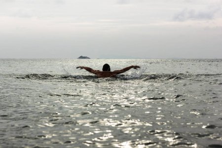 Téléchargez les photos : Silhouette de jeune homme nageant sur la technique du coup de papillon vers une petite île en arrière-plan au coucher du soleil à Koh Phangan, Thaïlande. Difficile à atteindre, défi de destination, concepts d'effort - en image libre de droit