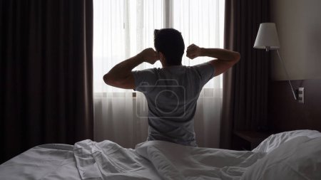 Photo for Silhouette of man sitting on bed stretching up arms by bedroom window. Early morning wake up, lazy, tired concepts - Royalty Free Image