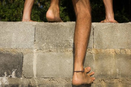 Téléchargez les photos : Gros plan sur les jambes d'un homme pieds nus escaladant un mur de briques. Immigration, concept d'intrusion - en image libre de droit