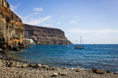 Téléchargez les photos : Superbes vues sur la plage de rochers par les falaises escarpées du littoral de Puerto de Mogan, Gran Canaria. Voilier sur une mer calme et paisible par une journée ensoleillée aux îles Canaries. Concept d'activités estivales en plein air - en image libre de droit