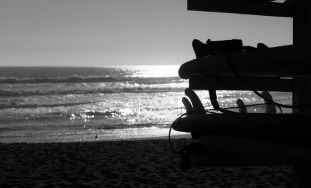 Foto de Silueta de tablas de surf tumbadas sobre soporte de madera con reflejo solar sobre el mar al atardecer en la playa de La Serena, Chile. Concepto de actividad deportiva de verano. Fotografía en blanco y negro - Imagen libre de derechos