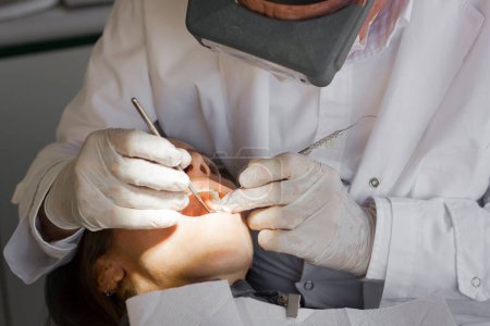 Foto de Dentist with special glasses and latex gloves using calculus plaque remover tooth scraper and mirror on female patient with mouth open. Health care, pain concept - Imagen libre de derechos