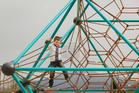 Little boy walking barefoot on red ropes while holding to pyramid net structure at playground on cloudy day in Bilbao. Child having fun in the park