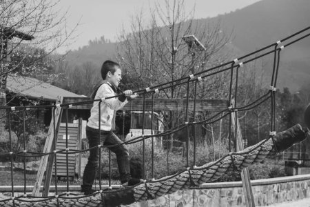 Téléchargez les photos : Jeune garçon traversant le pont tout en tenant des cordes au parc à Bilbao. Photographie noir et blanc. Enfant à l'aire de jeux - en image libre de droit