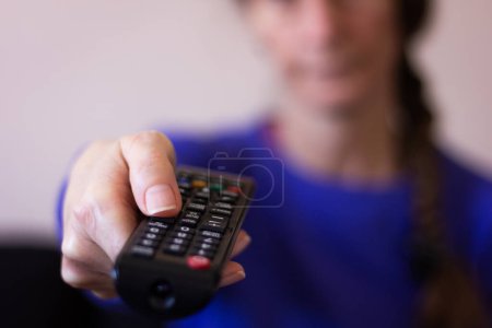 Téléchargez les photos : Gros plan de la main de la femme tenant télécommande tv et appuyant sur le bouton avec son visage flou sur le fond. Rester à la maison, divertissement, changer de concept de canaux - en image libre de droit