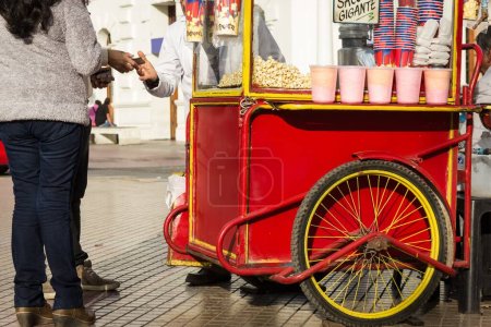 Téléchargez les photos : Homme et femme achetant du pop-corn sur une vieille charrette rouge pop-corn dans la rue avec des barbe à papa rose dans des verres en plastique à Plaza de Armas de La Serena, Chili. - en image libre de droit