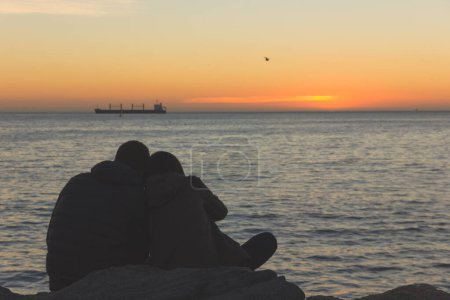 Téléchargez les photos : Silhouette de couple romantique assis sur des rochers tout en profitant du coucher de soleil avec cargo à l'horizon dans Via del Mar, Chili. Les amoureux se détendent au crépuscule - en image libre de droit