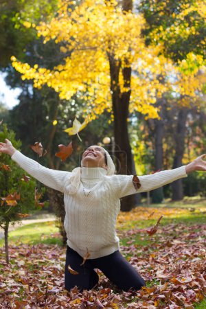 Photo for Happy mature woman on white turtle neck sweater and shawl covering her head enjoying fall season at park with open arms, knees on the ground of brown leaves and green and yellow trees in the forest - Royalty Free Image