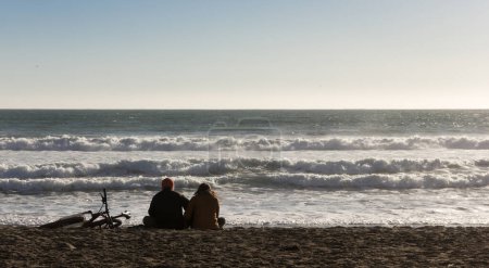 Foto de Silueta de pareja sentada sobre arena junto a bicicleta frente al mar agitado al atardecer en la playa de La Serena, Chile - Imagen libre de derechos