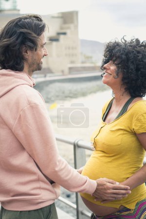 Foto de Pareja feliz esperando un bebé en un paseo por la playa. Mujer embarazada con camisa amarilla y padre sonriente mirándola el día ventoso en Las Palmas, España - Imagen libre de derechos