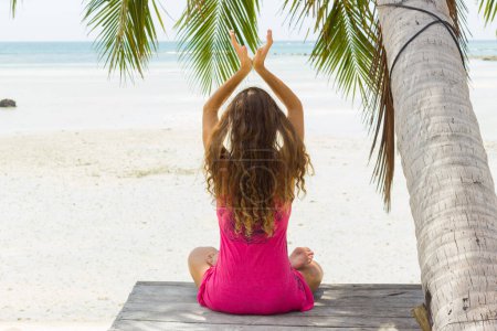 Téléchargez les photos : Pose de méditation, femme assise dans la pose de lotus sur la plage de sable sous le palmier à l'océan - en image libre de droit