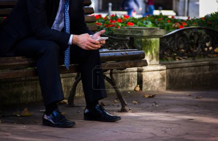 Photo for Businessman on black suit, shiny shoes and blue tie using smartphone while sitting on park bench in downtown Bilbao city. Full time workaholic concept. Stressed man touching cellphone screen - Royalty Free Image