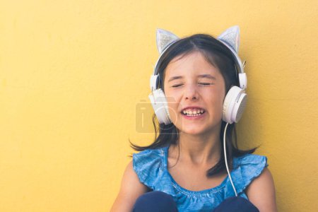 Photo for Little girl singing with funny design headphones on. Cute kid listening to music over yellow wall background. Pop star wannabe, fun, joy, happy child concepts - Royalty Free Image