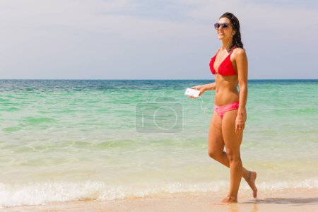 Foto de Chica feliz en bikini rojo y rosa caminando por la orilla del mar con gafas de sol y auriculares conectados a un móvil en la isla de Koh Pha Ngan, Tailandia. Vacaciones de verano, conceptos tecnológicos - Imagen libre de derechos
