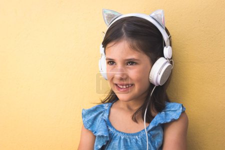 Téléchargez les photos : Petite fille souriante assise avec des écouteurs design d'oreilles de chat. Enfant joyeux avec t-shirt bleu sur fond mural jaune. Pop star aspirant, amusant, joie, technologie, concepts enfants heureux - en image libre de droit