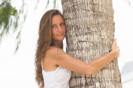 Foto de Retrato de mujer atractiva en vestido blanco y cabello largo y rizado abrazando una palmera en la playa en Tailandia. Sensual modelo femenina joven en el paraíso tropical. Exótico concepto de vacaciones de verano - Imagen libre de derechos