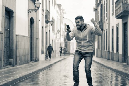 Photo for Exultant young man in the middle of road on rainy day feeling overexcited while looking at smart phone. Hipster gone crazy outdoors in the city. Sepia color - Royalty Free Image